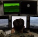 A military personnel member in camouflage uniform is seated at a multi-monitor workstation, appearing to operate a surveillance or targeting simulation system. The setup includes five NEC monitors displaying various feeds including a green night-vision-style view, daylight camera views of a mountainous terrain with structures, and a text interface. A logo reading 'BTS' is visible on one of the upper monitors, suggesting a training or simulation system.