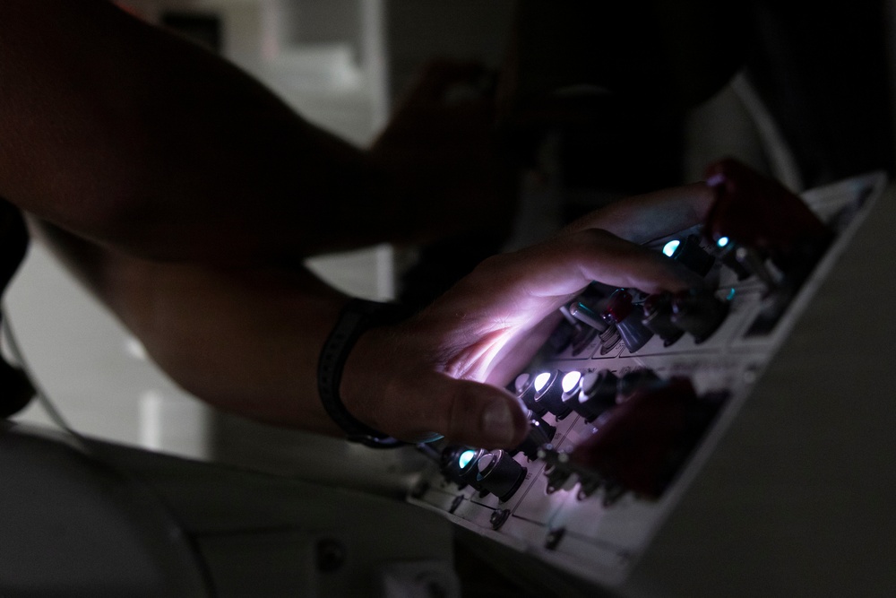A person's hands are operating or interacting with an illuminated control panel or electronic equipment in a darkened environment. The panel features multiple buttons and indicator lights that are actively glowing, casting light onto the operator's hands. The low-light setting emphasizes the luminous controls and creates a dramatic, high-contrast composition.