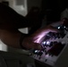 A person's hands are operating or interacting with an illuminated control panel or electronic equipment in a darkened environment. The panel features multiple buttons and indicator lights that are actively glowing, casting light onto the operator's hands. The low-light setting emphasizes the luminous controls and creates a dramatic, high-contrast composition.