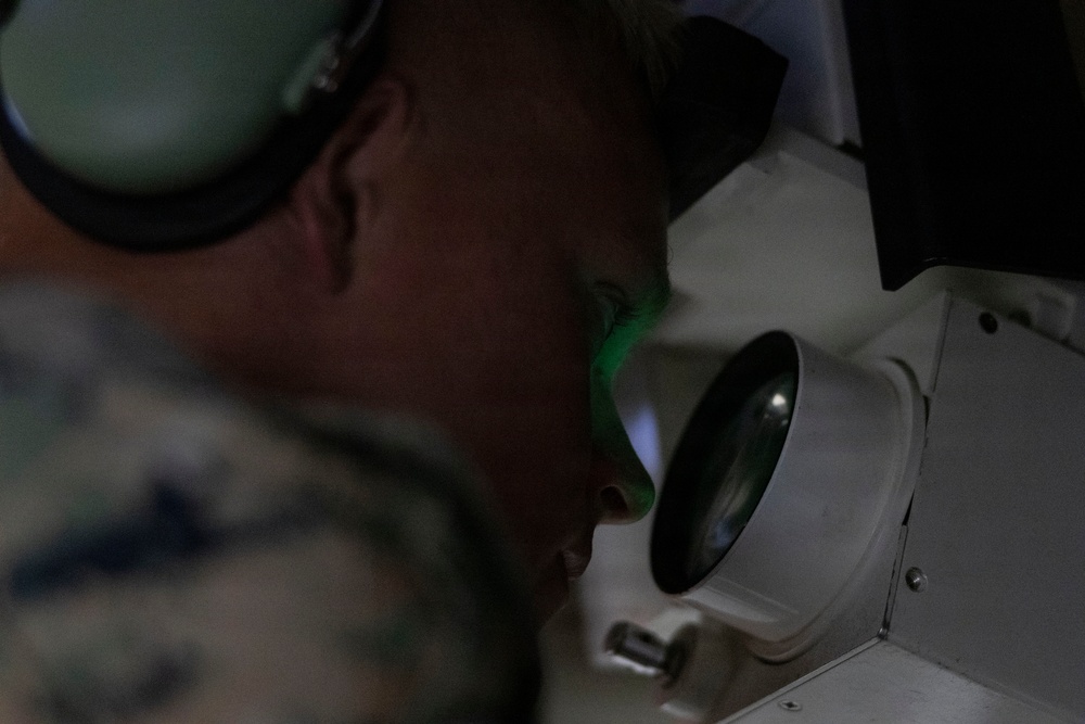 A military personnel member wearing camouflage uniform and hearing protection (ear muffs) is looking through a large optical lens or sight device. A green light is visible reflecting on their face from the optic. The image is taken in close-up, low-light conditions emphasizing the optical equipment.