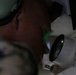 A military personnel member wearing camouflage uniform and hearing protection (ear muffs) is looking through a large optical lens or sight device. A green light is visible reflecting on their face from the optic. The image is taken in close-up, low-light conditions emphasizing the optical equipment.