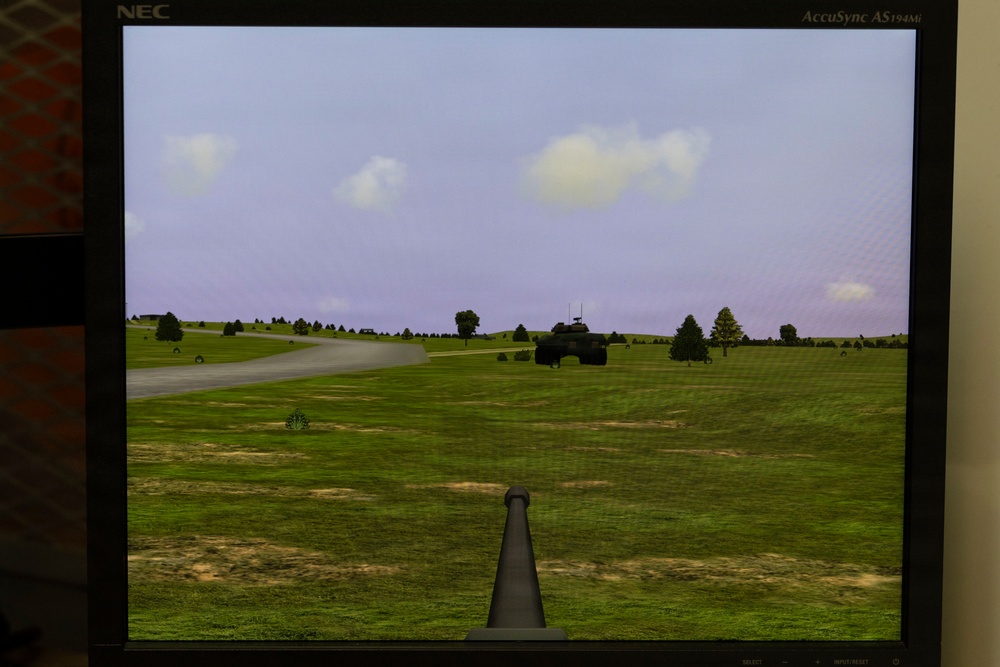 A photograph of an NEC AccuSync AS194Mi monitor displaying a military vehicle simulation or video game. The screen shows a first-person perspective from what appears to be a tank or armored vehicle, with a gun barrel visible in the foreground and an enemy or allied armored vehicle in the mid-distance. The virtual environment features a green grassy landscape, a road, scattered trees, and a partly cloudy sky.