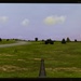 A photograph of an NEC AccuSync AS194Mi monitor displaying a military vehicle simulation or video game. The screen shows a first-person perspective from what appears to be a tank or armored vehicle, with a gun barrel visible in the foreground and an enemy or allied armored vehicle in the mid-distance. The virtual environment features a green grassy landscape, a road, scattered trees, and a partly cloudy sky.