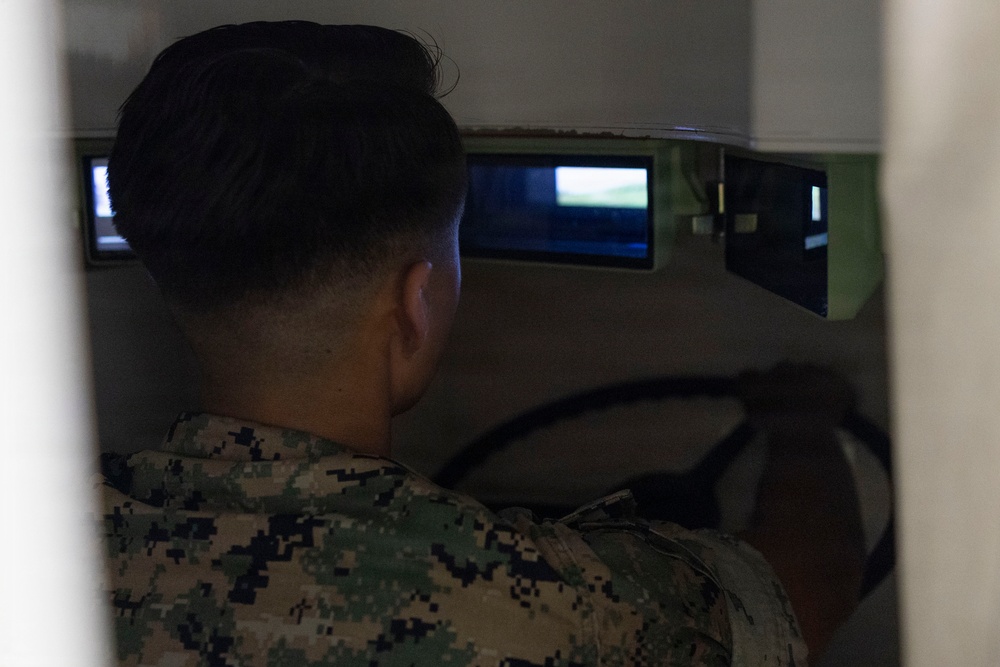 A military service member in digital camouflage uniform is seated at what appears to be a vehicle or driving simulator, viewed from behind. Multiple display screens are visible in front of the individual, suggesting a simulation training environment. The image is taken from outside the simulator enclosure, partially obscured by a structural element.