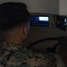 A military service member in digital camouflage uniform is seated at what appears to be a vehicle or driving simulator, viewed from behind. Multiple display screens are visible in front of the individual, suggesting a simulation training environment. The image is taken from outside the simulator enclosure, partially obscured by a structural element.