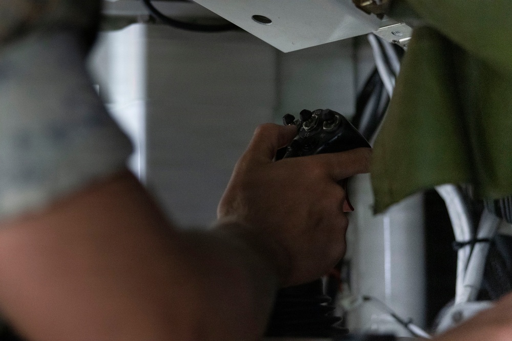 A person is working with their hands on what appears to be electrical or mechanical equipment in a confined space. They are holding a dark-colored tool or connector component and appear to be installing or adjusting it on an overhead panel or surface. The image is taken at close range with significant depth-of-field blur on the foreground elements.