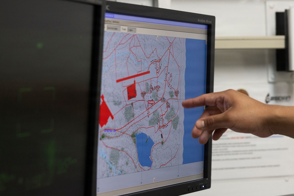 A person is pointing at a computer monitor displaying a digital map with red overlay markings, likely a GIS or tactical mapping application. The map shows terrain features including water bodies (shown in blue), roads or boundaries (shown in red lines), and various geographic annotations. The setting appears to be an office or operations center with multiple monitors visible.