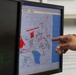 A person is pointing at a computer monitor displaying a digital map with red overlay markings, likely a GIS or tactical mapping application. The map shows terrain features including water bodies (shown in blue), roads or boundaries (shown in red lines), and various geographic annotations. The setting appears to be an office or operations center with multiple monitors visible.