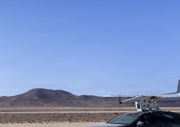 A dark-colored sedan, appearing to be a Toyota, is driving across a dry desert lakebed with a fixed-wing UAV (unmanned aerial vehicle) mounted on a roof rack. The vehicle is kicking up dust as it moves, suggesting it is being used as a mobile launch platform for the drone. The setting appears to be a remote arid desert environment with low hills visible in the background.