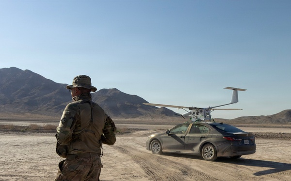 A military personnel in camouflage uniform and boonie hat stands on a dry desert lakebed observing a civilian sedan that has a fixed-wing UAV (unmanned aerial vehicle) mounted on a launch platform on its roof. The vehicle appears to be accelerating along a dirt track, likely to generate sufficient speed for drone launch. The setting appears to be an arid desert testing or training environment with mountain ranges visible in the background.