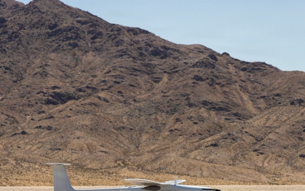 A white fixed-wing unmanned aerial vehicle (UAV) or small aircraft is captured at low altitude over a dry desert lakebed, appearing to be either landing or taking off. The arid, flat terrain is characteristic of a dry lake or playa, with rugged, rocky desert mountains visible in the background. The scene suggests a remote test or operational flight environment.