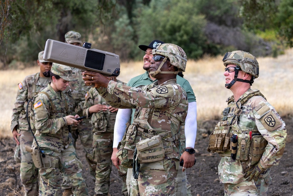 A group of U.S. Army soldiers in OCP (Operational Camouflage Pattern) uniforms are gathered outdoors in a dry, wooded training area. A central soldier is holding up what appears to be a counter-drone or directed-energy device, possibly a drone detection/jamming system, while others observe. The scene appears to be a field demonstration or training exercise.