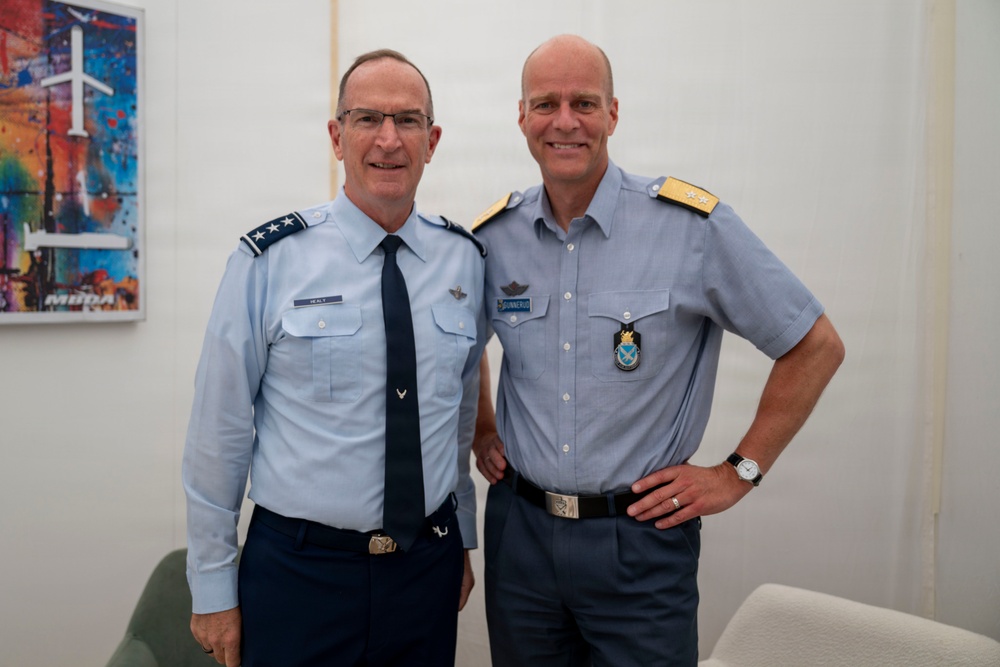 Two senior military officers in light blue uniforms pose together for a photograph indoors. The officer on the left wears a US Air Force uniform with three-star rank insignia (Lieutenant General), while the officer on the right wears what appears to be a Scandinavian/Nordic air force uniform with two-star shoulder boards and a distinctive badge. Both are smiling in what appears to be an official or diplomatic meeting setting.