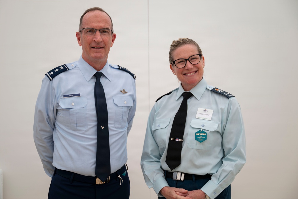 Two military officers in uniform are posing together for a photograph against a plain light-colored background. The officer on the left wears a US Air Force uniform with two-star (Major General) rank insignia and a name tag reading 'HEALY'. The officer on the right wears what appears to be a Royal Australian Air Force uniform with an Air Commodore rank insignia and a name badge/event tag.