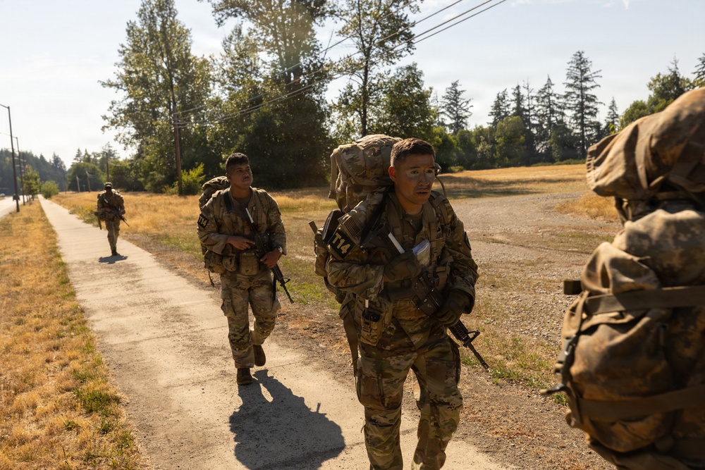 A group of soldiers in full combat gear and large rucksacks are conducting a road march along a paved path in what appears to be a military training area. The soldiers are wearing OCP (Operational Camouflage Pattern) uniforms and carrying rifles, with visible numbered identification tags on their chest. The setting appears to be a sunny day in a Pacific Northwest-style environment with dry grass fields and coniferous trees.
