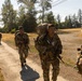 A group of soldiers in full combat gear and large rucksacks are conducting a road march along a paved path in what appears to be a military training area. The soldiers are wearing OCP (Operational Camouflage Pattern) uniforms and carrying rifles, with visible numbered identification tags on their chest. The setting appears to be a sunny day in a Pacific Northwest-style environment with dry grass fields and coniferous trees.