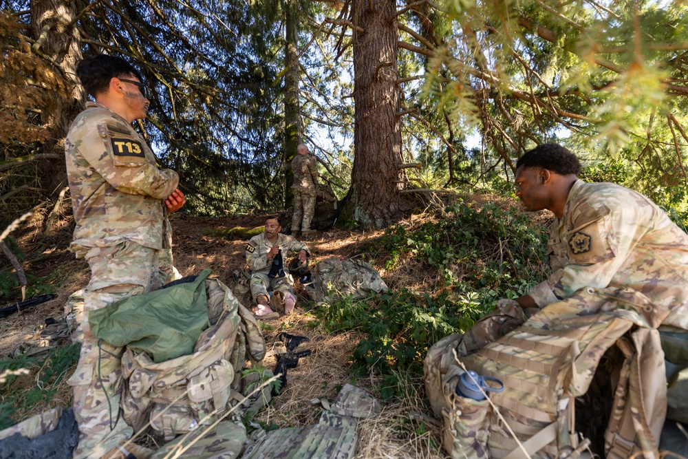 A group of military personnel in MultiCam/OCP camouflage uniforms are gathered in a wooded area, appearing to be on a rest break or conducting a field training exercise. At least four soldiers are visible among large conifer trees, with tactical gear and rucksacks scattered around them. One soldier on the left displays a 'T13' identifier patch, suggesting a training or competition event.