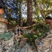 A group of military personnel in MultiCam/OCP camouflage uniforms are gathered in a wooded area, appearing to be on a rest break or conducting a field training exercise. At least four soldiers are visible among large conifer trees, with tactical gear and rucksacks scattered around them. One soldier on the left displays a 'T13' identifier patch, suggesting a training or competition event.