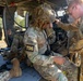 U.S. Army soldiers are conducting what appears to be a pre-jump or equipment check inside a military helicopter, likely during airborne or air assault training. A soldier marked 'T06' is seated on the helicopter floor while another soldier assists with gear/harness adjustments. The setting appears to be a training exercise at a military installation surrounded by forested terrain.