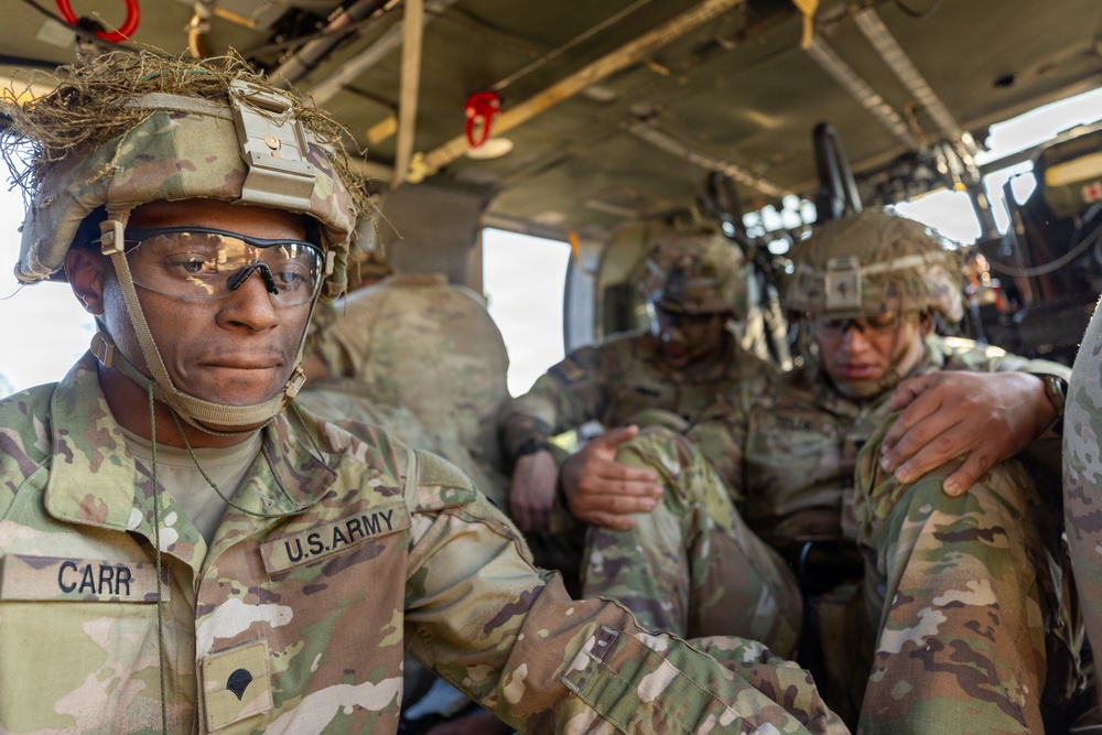 Several U.S. Army soldiers in full combat gear are seated inside what appears to be a military helicopter or transport vehicle. The soldier in the foreground, identified by name tape as 'CARR,' wears OCP (Operational Camouflage Pattern) uniform, a tactical helmet with camouflage netting, and ballistic eyewear. The soldiers appear to be in transit, likely during a training exercise or operational deployment.