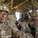Several U.S. Army soldiers in full combat gear are seated inside what appears to be a military helicopter or transport vehicle. The soldier in the foreground, identified by name tape as 'CARR,' wears OCP (Operational Camouflage Pattern) uniform, a tactical helmet with camouflage netting, and ballistic eyewear. The soldiers appear to be in transit, likely during a training exercise or operational deployment.