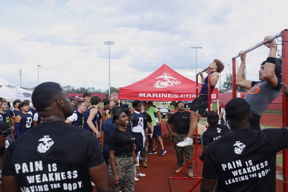 A U.S. Marines recruiting event is taking place at an outdoor athletic track facility. Young athletes, likely high school football players, are participating in a pull-up challenge on a red pull-up bar station while Marine recruiters in black 'Pain is Weakness Leaving the Body' t-shirts observe and supervise. A red Marines.com branded canopy tent is visible in the background.