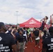 A U.S. Marines recruiting event is taking place at an outdoor athletic track facility. Young athletes, likely high school football players, are participating in a pull-up challenge on a red pull-up bar station while Marine recruiters in black 'Pain is Weakness Leaving the Body' t-shirts observe and supervise. A red Marines.com branded canopy tent is visible in the background.