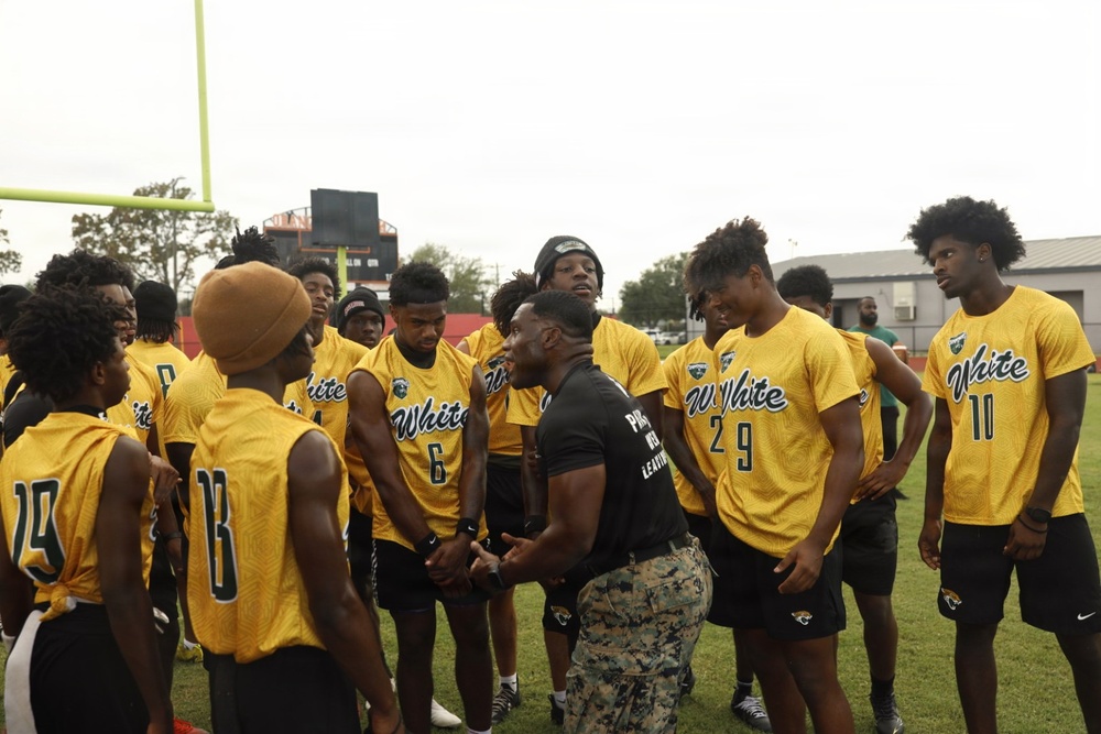 A group of young football players wearing yellow 'White' jerseys are gathered in a huddle on a football field. A man in a black shirt and camouflage pants, appearing to be a coach or instructor, is addressing the team in the center of the group. The setting appears to be an outdoor football field with goal posts and stadium structures visible in the background.
