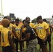 A group of young football players wearing yellow 'White' jerseys are gathered in a huddle on a football field. A man in a black shirt and camouflage pants, appearing to be a coach or instructor, is addressing the team in the center of the group. The setting appears to be an outdoor football field with goal posts and stadium structures visible in the background.