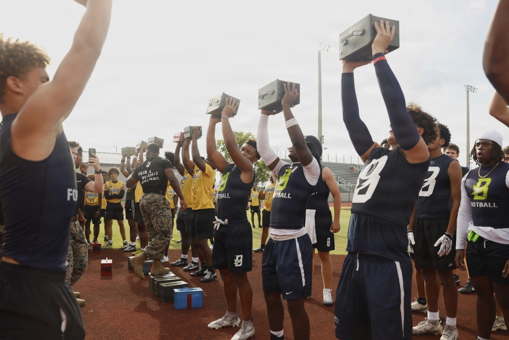 A group of high school football players in navy blue and gold uniforms are participating in a military-style fitness challenge on a track field, lifting what appear to be military ammunition cans overhead. A person in Marine Corps camouflage uniform is visible among the participants, suggesting this is a Marine Corps-sponsored athletic event or combine. The activity appears to be a timed overhead press or endurance hold exercise.
