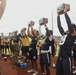 A group of high school football players in navy blue and gold uniforms are participating in a military-style fitness challenge on a track field, lifting what appear to be military ammunition cans overhead. A person in Marine Corps camouflage uniform is visible among the participants, suggesting this is a Marine Corps-sponsored athletic event or combine. The activity appears to be a timed overhead press or endurance hold exercise.