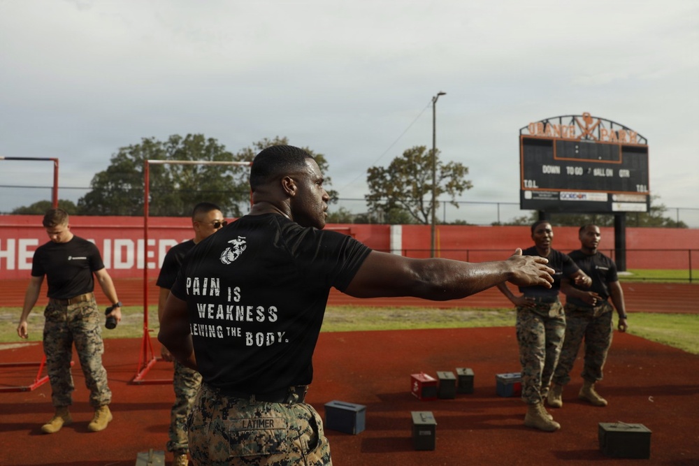 A group of U.S. Marines are conducting a physical training exercise on a high school football field track. The central figure, identified by name tape as 'LATIMER,' wears a black t-shirt reading 'PAIN IS WEAKNESS LEAVING THE BODY' with the USMC Eagle, Globe, and Anchor emblem. Several other Marines in camouflage utilities are visible in the background near what appear to be ammunition cans used as exercise equipment.
