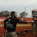 A group of U.S. Marines are conducting a physical training exercise on a high school football field track. The central figure, identified by name tape as 'LATIMER,' wears a black t-shirt reading 'PAIN IS WEAKNESS LEAVING THE BODY' with the USMC Eagle, Globe, and Anchor emblem. Several other Marines in camouflage utilities are visible in the background near what appear to be ammunition cans used as exercise equipment.