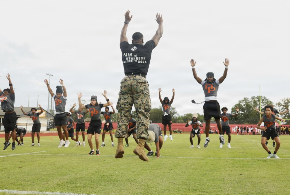 A U.S. Marine in camouflage utilities and tan boots leads a group of youth football players in jumping jacks or a similar calisthenics exercise on a grass athletic field. The Marine's black t-shirt reads 'PAIN IS WEAKNESS LEAVING THE BODY' with the Marine Corps Eagle, Globe, and Anchor emblem. The athletes are wearing matching gray and black uniforms with an orange 'DP' logo, suggesting a school or team identity.