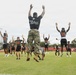 A U.S. Marine in camouflage utilities and tan boots leads a group of youth football players in jumping jacks or a similar calisthenics exercise on a grass athletic field. The Marine's black t-shirt reads 'PAIN IS WEAKNESS LEAVING THE BODY' with the Marine Corps Eagle, Globe, and Anchor emblem. The athletes are wearing matching gray and black uniforms with an orange 'DP' logo, suggesting a school or team identity.