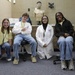 Four young people pose together in what appears to be a medical or science laboratory setting. Two of them are wearing white lab coats, while the others wear casual yellow t-shirts with lanyards. A medical training mannequin torso is visible in the background.