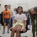 A U.S. Army soldier in camouflage uniform interacts with a group of children, appearing to demonstrate something to a girl seated in an office chair. The setting looks like an indoor facility, possibly a military base during an educational or community outreach event. Several children of varying ages are observing the interaction with interest.