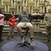 A group of children and a U.S. Army soldier are inside what appears to be an anechoic chamber, identifiable by the distinctive sound-absorbing foam wedge panels covering the walls. A teenage boy is seated in a specialized rotating or positioning chair on a raised platform, with his arms outstretched and eyes closed, appearing to participate in a demonstration or experiment. A U.S. Army soldier holding a clipboard observes alongside two other children.