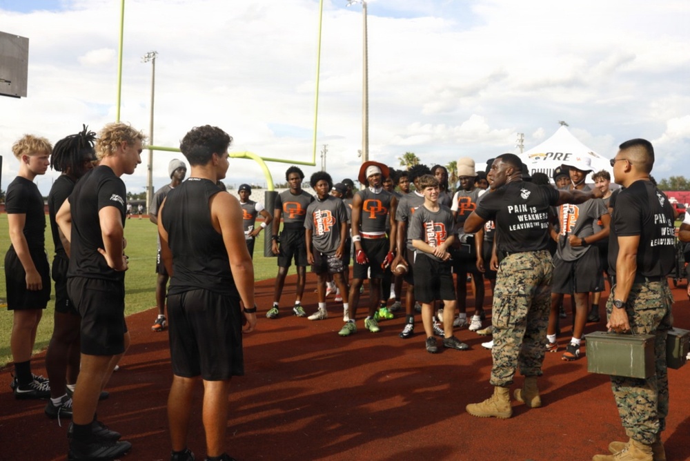 Two U.S. Marines in camouflage pants and black 'Pain is Weakness Leaving the Body' shirts are addressing a large group of high school football players on a track and field facility. The football players are wearing gray jerseys with an orange 'P' logo, suggesting they represent a school team. One Marine appears to be speaking or presenting to the assembled athletes, possibly as part of a recruiting or motivational event.