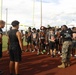 Two U.S. Marines in camouflage pants and black 'Pain is Weakness Leaving the Body' shirts are addressing a large group of high school football players on a track and field facility. The football players are wearing gray jerseys with an orange 'P' logo, suggesting they represent a school team. One Marine appears to be speaking or presenting to the assembled athletes, possibly as part of a recruiting or motivational event.