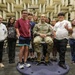 A U.S. Army soldier in camouflage uniform sits in a chair on a rotating platform surrounded by a group of approximately eight middle-school-aged children. The group is posed inside what appears to be an anechoic chamber, identifiable by the distinctive sound-absorbing foam wedge panels covering the walls. This appears to be an educational or STEM outreach visit to a military or research facility.