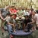 A U.S. Army soldier in camouflage uniform sits on a stool surrounded by a group of approximately 11 children and young teenagers posing for a group photo. They are inside an anechoic chamber, identifiable by the distinctive foam acoustic wedge panels covering the walls and ceiling. The setting suggests an educational tour or STEM outreach event at a military or research facility.