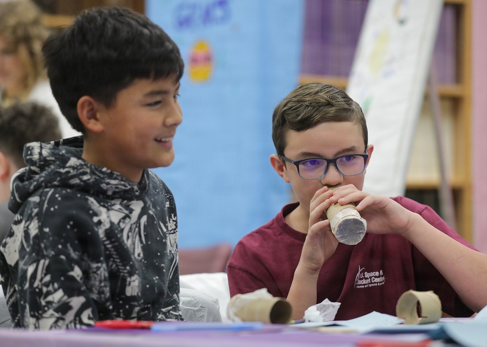 Two young boys are engaged in a hands-on classroom activity, likely a STEM or science project. The boy on the right, wearing glasses and a U.S. Space &amp; Rocket Center t-shirt, is blowing into a cardboard tube, while the boy on the left watches with a smile. The setting appears to be a school classroom with educational materials visible in the background.
