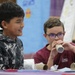 Two young boys are engaged in a hands-on classroom activity, likely a STEM or science project. The boy on the right, wearing glasses and a U.S. Space &amp; Rocket Center t-shirt, is blowing into a cardboard tube, while the boy on the left watches with a smile. The setting appears to be a school classroom with educational materials visible in the background.