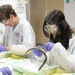 Two students in a laboratory setting are performing what appears to be a dissection activity, likely on eels or similar elongated specimens. Both are wearing standard lab safety equipment including white lab coats, purple nitrile gloves, and safety goggles. The setting appears to be a school or educational science lab.