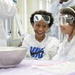 Two young children are participating in a science experiment or educational activity, wearing white lab coats and safety goggles. A metal mixing bowl is visible in the foreground, possibly containing a substance producing vapor or foam. The children appear excited and engaged, suggesting a hands-on STEM learning environment.