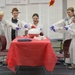 Five children dressed in white lab coats, safety goggles, and purple gloves are participating in a science demonstration in what appears to be a classroom setting. They are gathered around a red-covered table where a large inflated balloon or dome-shaped object is the focal point of their experiment. The children are excitedly pointing at the object, suggesting a successful or surprising result from their activity.