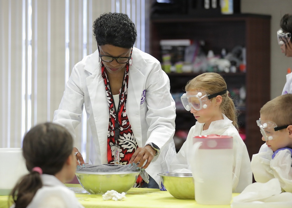 A science educator in a white lab coat and glasses leads a hands-on science experiment with young children at a yellow-covered table. The children are wearing white lab coats and protective safety goggles, indicating a structured educational science activity. The setting appears to be a classroom or science education facility with supply shelves visible in the background.