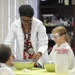 A science educator in a white lab coat and glasses leads a hands-on science experiment with young children at a yellow-covered table. The children are wearing white lab coats and protective safety goggles, indicating a structured educational science activity. The setting appears to be a classroom or science education facility with supply shelves visible in the background.