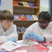 Two young students are engaged in a hands-on science activity, wearing white lab coats, blue nitrile gloves, and safety glasses while examining what appear to be biological or geological specimens. They are seated at a red-covered table in what appears to be a library or learning center, with reference materials and books visible in the background. The student on the right is actively handling a specimen while the student on the left observes closely.